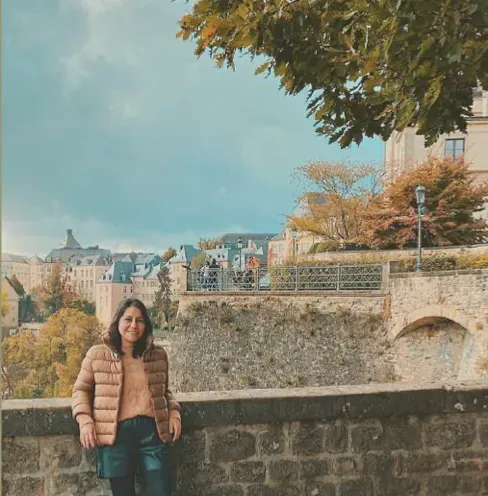 Lydia adossée à un muret de pierre, une vieille ville et des arbres d’automne derrière elle